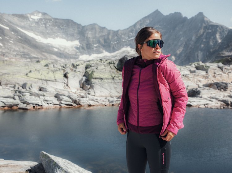 Frau in pinker Outdoorjacke und Sportbrille vor einem Bergsee in alpiner Landschaft.