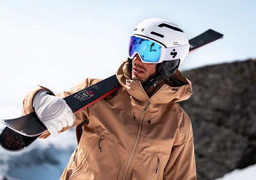 Skifahrer mit Helm und Skibrille trägt seine Ski über der Schulter im verschneiten Hochgebirge.