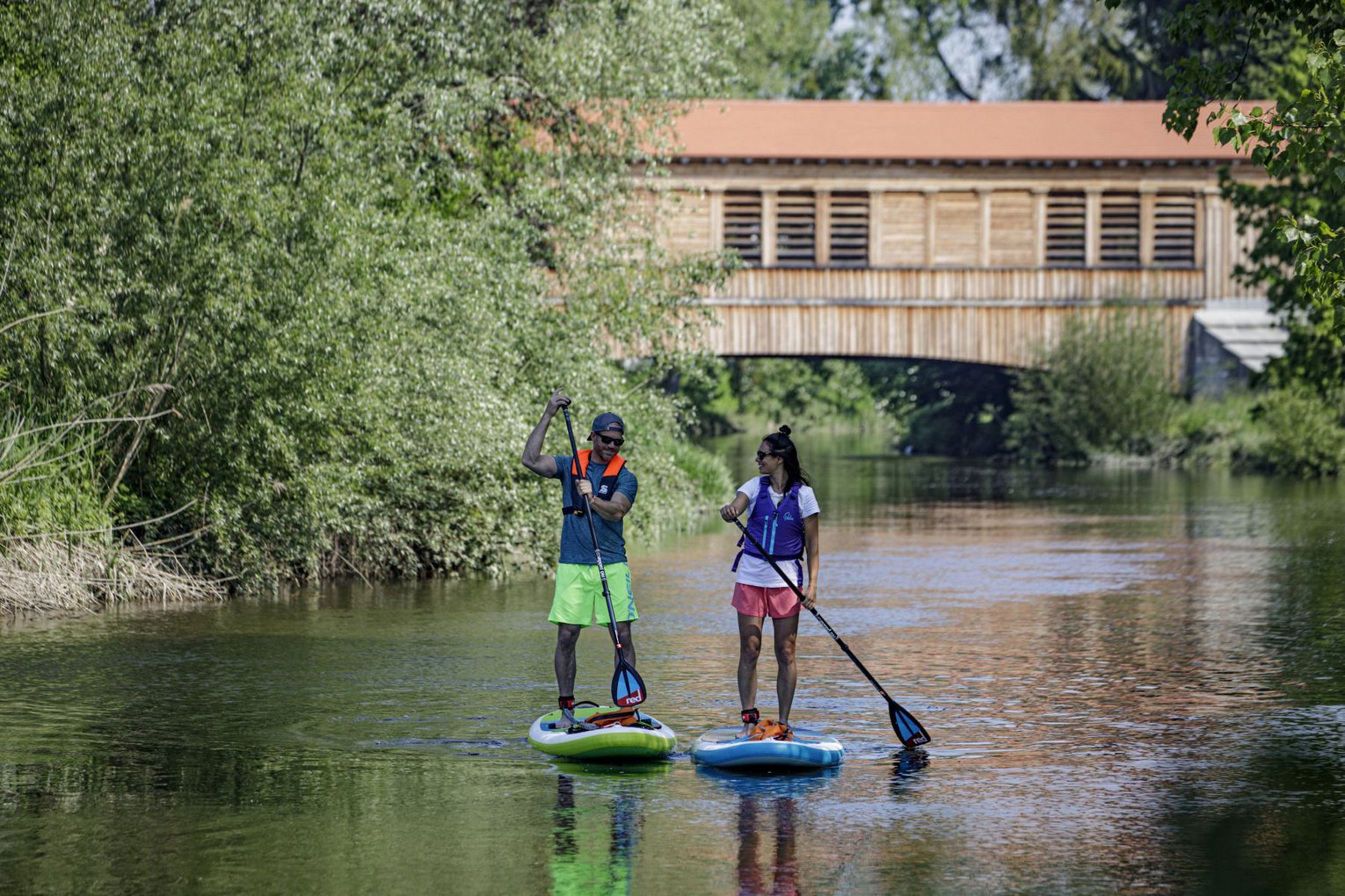 Wassersport Zwei Personen paddeln mit SUP auf einem Fluß, im Hintergrund eine Holzbrücke